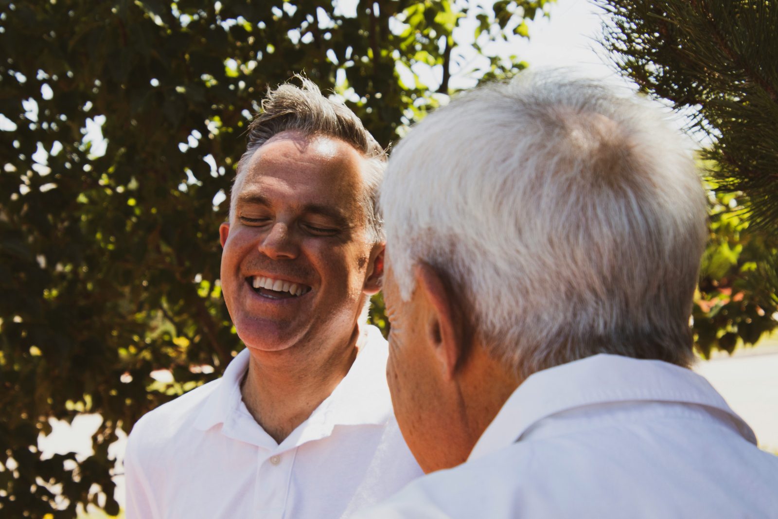 Two men are seen outdoors, engaged in a conversation. The man on the left is smiling with his eyes closed, while facing the man on the right who has his back to the camera. Both are wearing white shirts, and trees and greenery can be seen in the background.