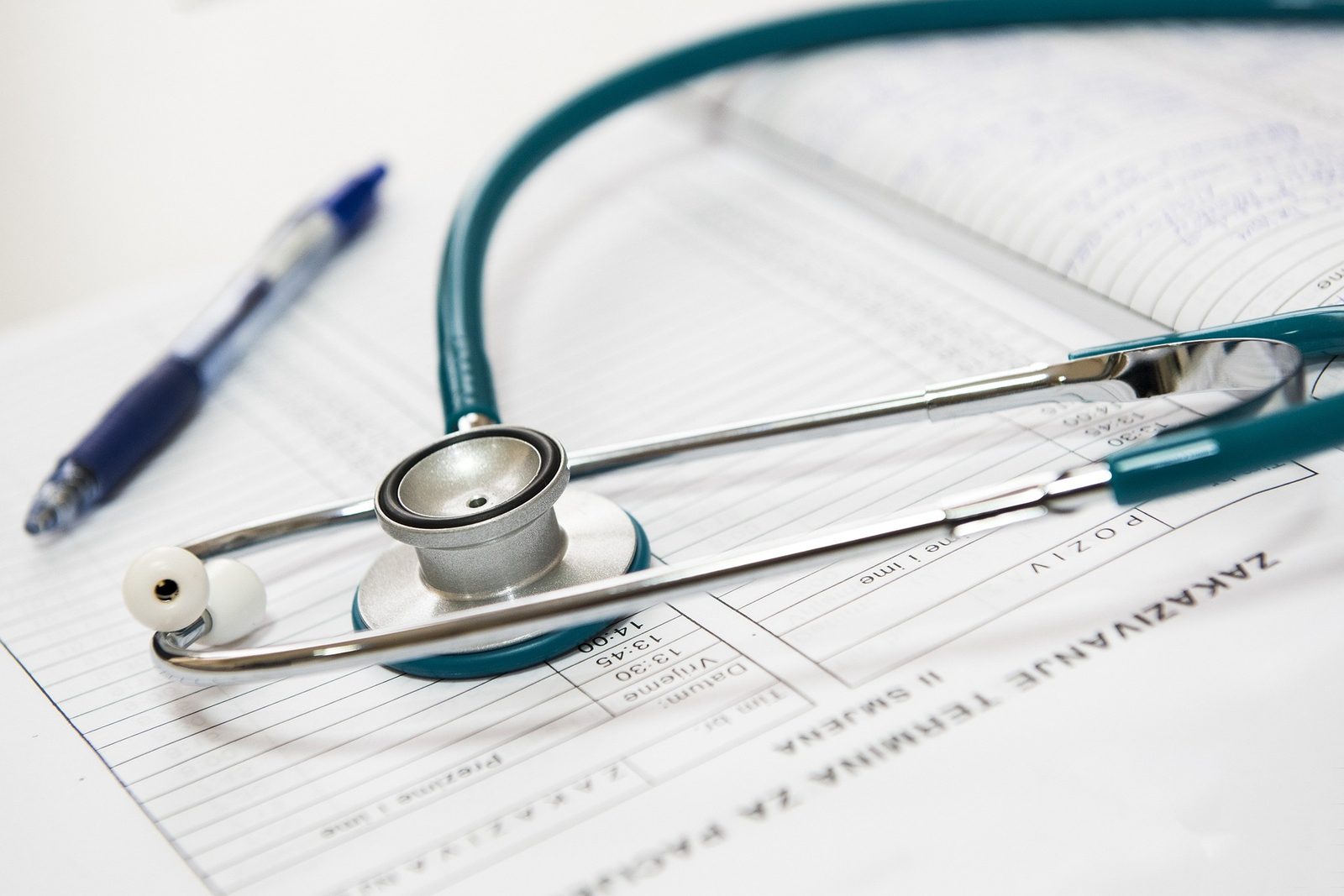 A stethoscope lies on a medical document that appears to be a patient chart or form. A blue pen is placed to the left of the stethoscope. The background shows blurred text, focusing attention on the medical tools.