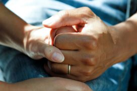 Close-up view of two hands clasped together. One hand is wearing a wedding ring and gently holds the other, which is resting on a fabric surface. The image conveys a sense of comfort and support.