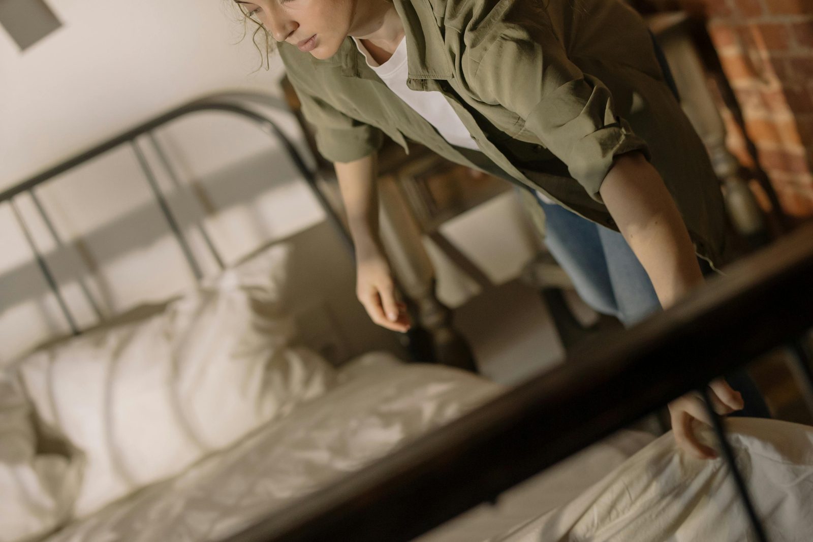 A person wearing a green jacket is making a bed with white sheets. The bed has a black metal frame, and the room features exposed brick walls and wooden furniture. Sunlight casts shadows through a window, creating a cozy and airy atmosphere.