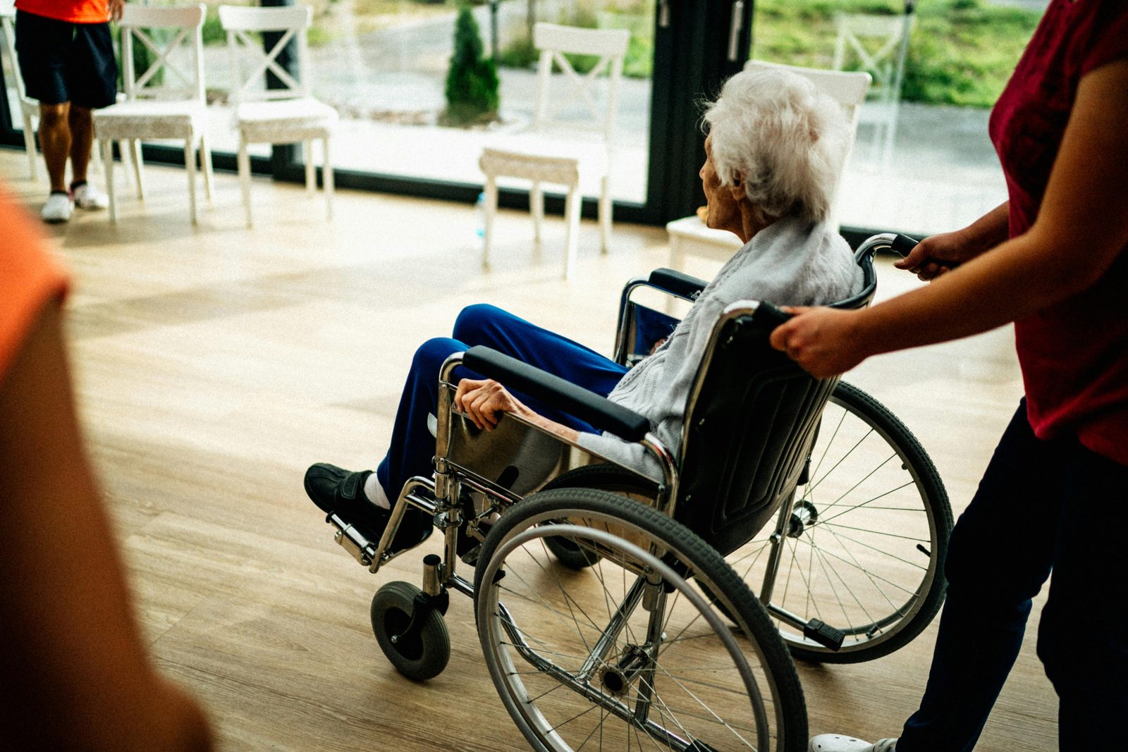 An elderly person with white hair is seated in a wheelchair, being pushed by another individual. They are indoors, with several empty chairs and large windows in the background, allowing natural light to fill the space.