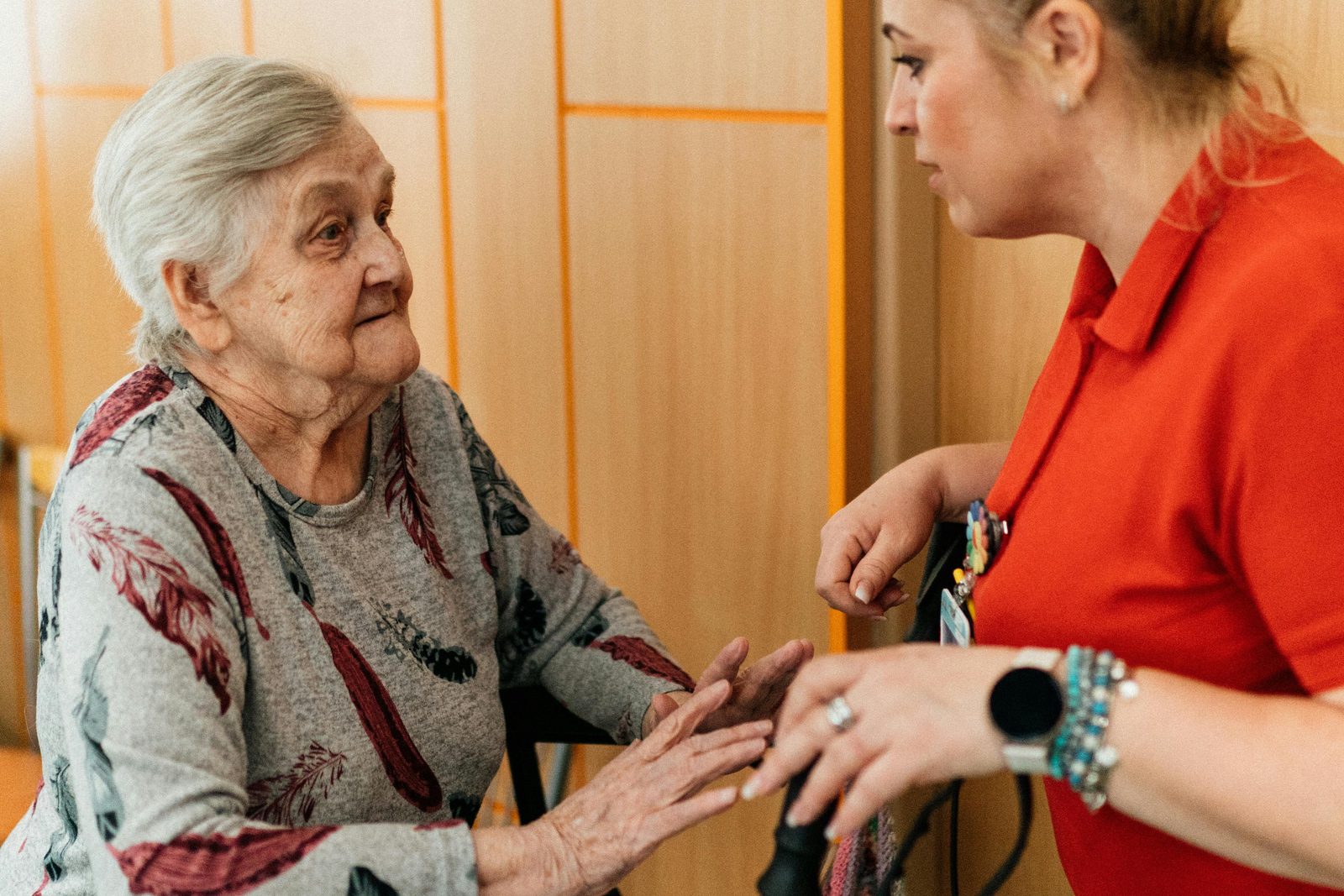 An elderly woman, seated, speaks with a caregiver in a red shirt. The caregiver is standing, facing her, holding her hands, and wearing a watch and bracelets. They are in a warmly lit room with wooden paneled walls.