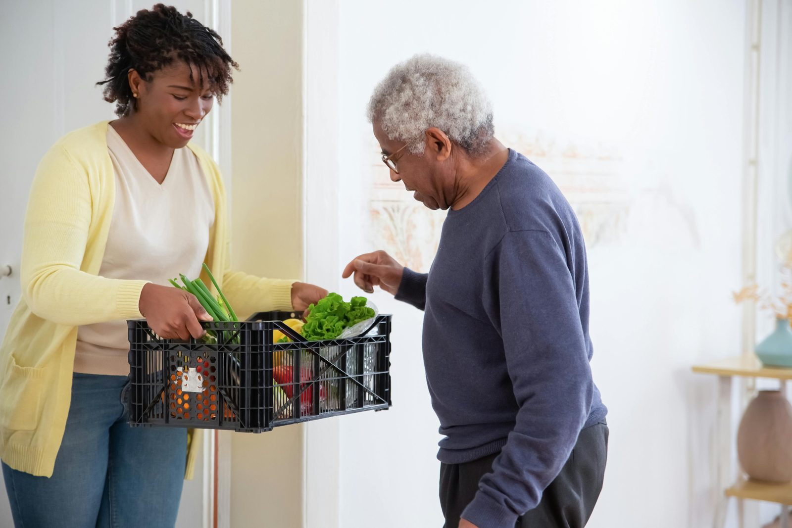 A young woman wearing a yellow cardigan hands a black crate of fresh vegetables to an elderly man with gray hair and glasses. They both appear to be smiling, and the man is wearing a dark blue sweater. They are standing in a bright, cozy room.