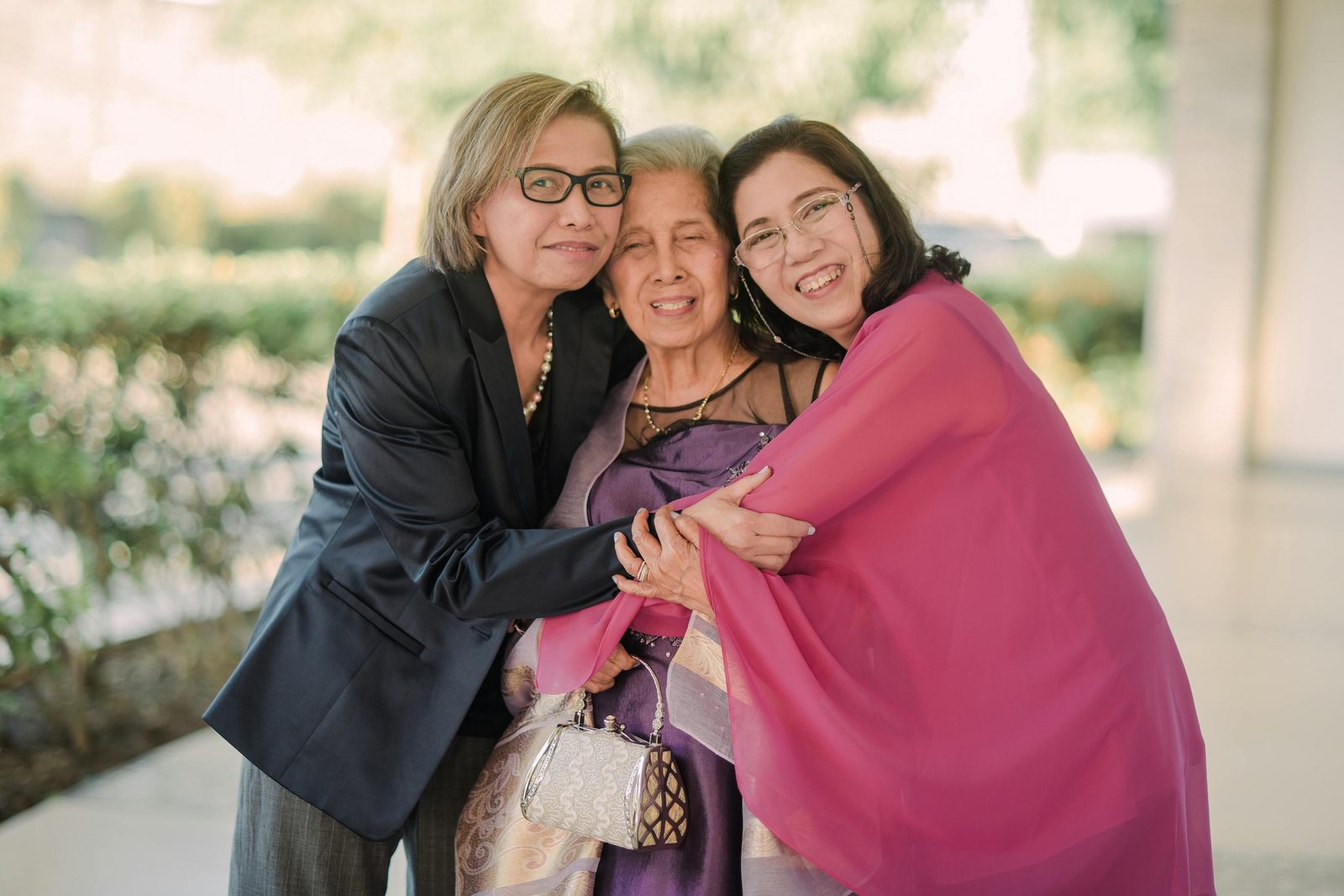 Three older women are standing outside, smiling and embracing. The woman in the middle is holding a small, textured handbag. All three are dressed in formal attire, with the women on the left and right wearing glasses. The background is blurred greenery.