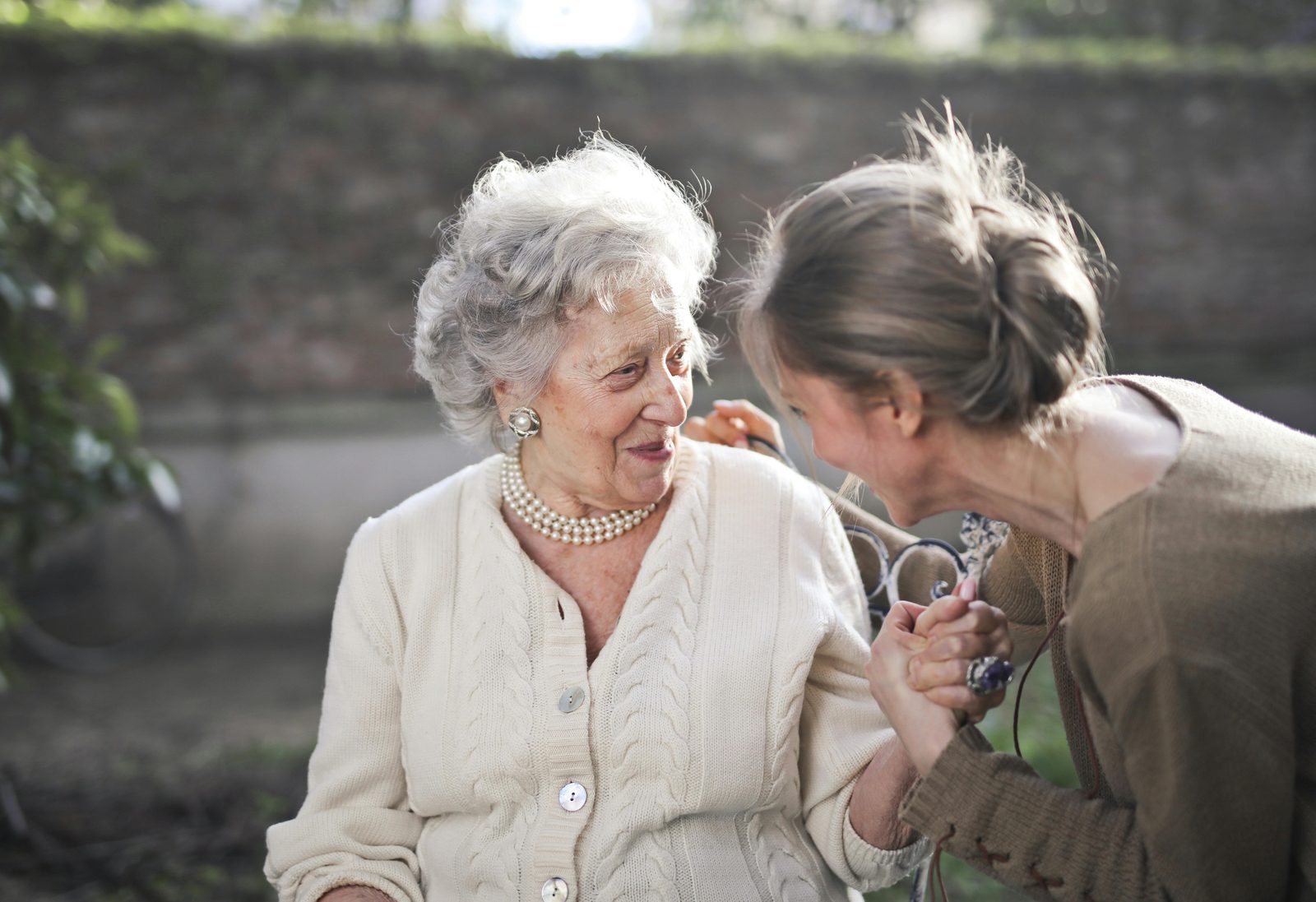 An elderly woman with gray hair, wearing a beige cardigan and pearl necklace, sits outside smiling and holding hands with a younger woman who is leaning towards her, also smiling. The background is a blurred garden scene with greenery and a brick wall.