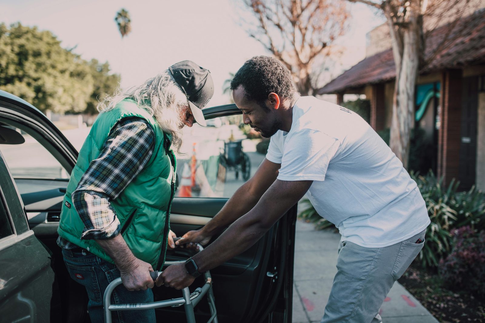 A young man is assisting an elderly person with white hair and a green vest in using a walker to exit a car. They are outside, near a sidewalk with trees and a building in the background. The young man is holding the walker, providing support.