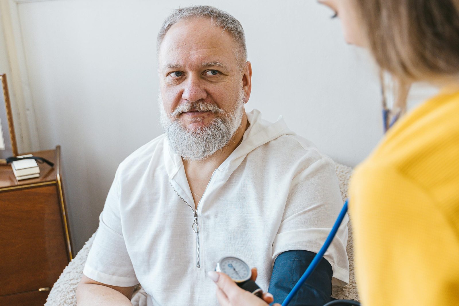 A bearded man in a white shirt is sitting down and having his blood pressure checked by a healthcare professional in a yellow top, who is holding a blood pressure cuff around his arm and a stethoscope. The setting appears to be a home or a cozy room.