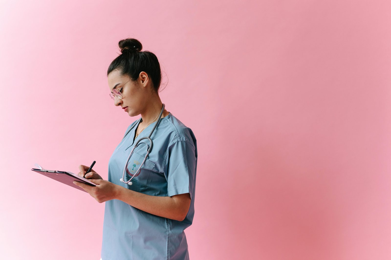 A nurse stands against a pink background, wearing light blue scrubs and a stethoscope around her neck. She is writing on a clipboard, focused on her task, with her dark hair tied up in a bun.